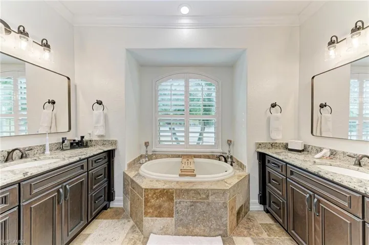 Full bath featuring stone tile flooring, two vanities, ornamental molding, and a garden tub