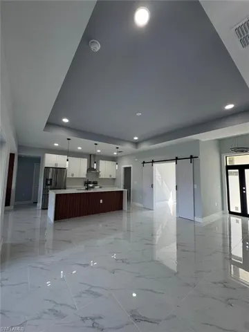 Kitchen with wall chimney range hood, stainless steel fridge, white cabinetry, hanging light fixtures, and an island with sink