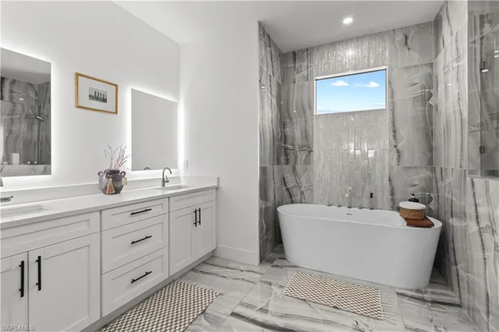 Bathroom with a marble finish shower, double vanity, light marble finish flooring, a freestanding tub, and tile walls