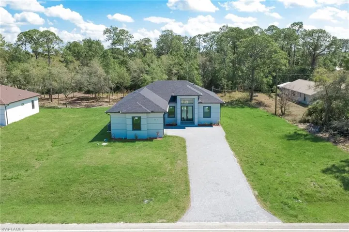 View of front facade with driveway, roof with shingles, a front lawn, and view of wooded area
