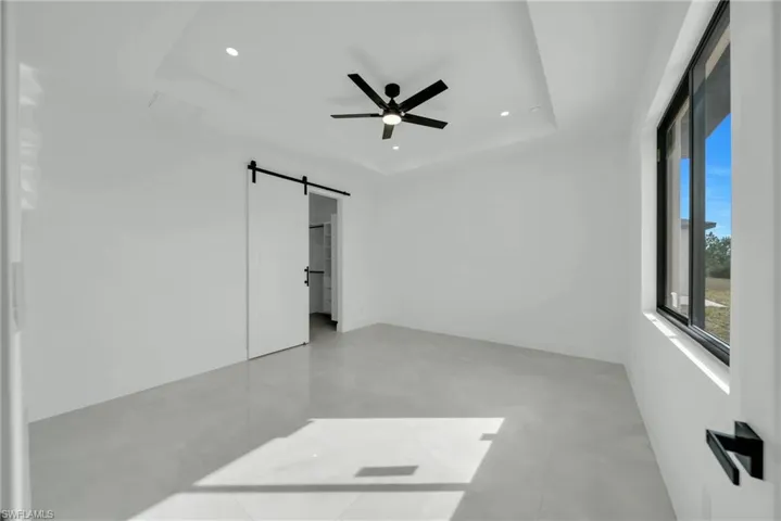 Unfurnished bedroom featuring a barn door, concrete floors, recessed lighting, ceiling fan, and a tray ceiling
