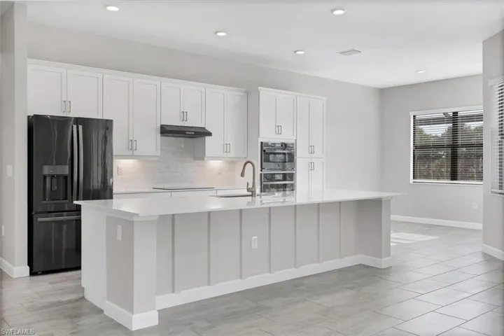 Kitchen with fridge with ice dispenser, double oven, an island with sink, light countertops, and under cabinet range hood