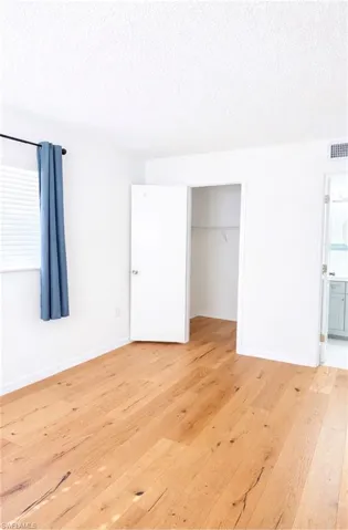 Unfurnished bedroom with a textured ceiling, light wood-style floors, a walk in closet, ensuite bath, and multiple windows