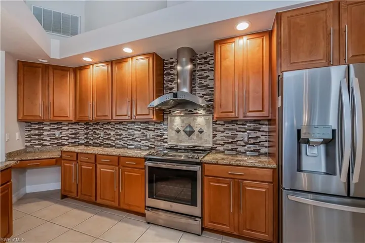 Kitchen featuring backsplash, wall chimney range hood, light tile patterned floors, light stone counters, and stainless steel appliances