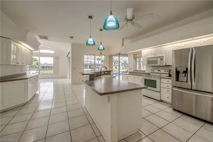 Kitchen featuring white appliances, light tile patterned floors, white cabinets, an island with sink, and hanging light fixtures