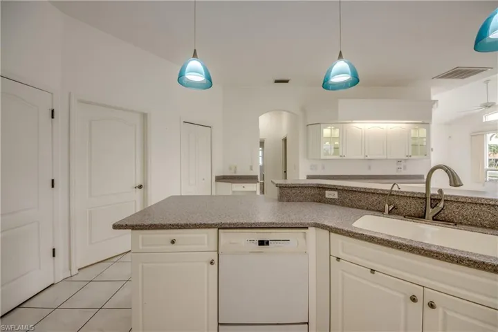 Kitchen featuring white cabinetry, light tile patterned floors, dishwasher, pendant lighting, and sink.  Entrance to Guest Wing beyond the arched doorway