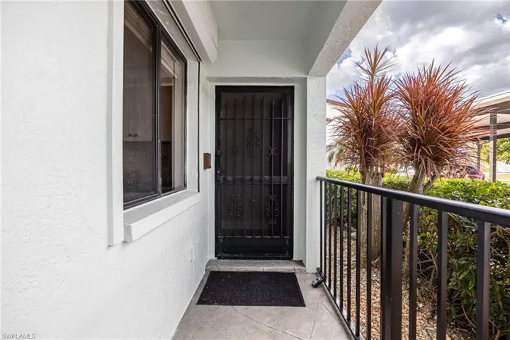 Entryway featuring a black security door, a fixed window with a roller shutter, white stucco exterior, black metal railing, and tropical landscaping