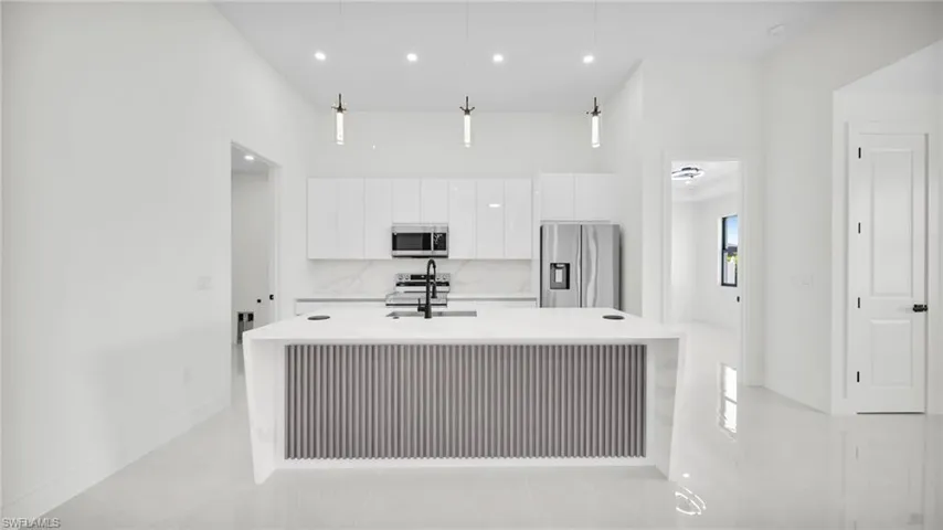 Kitchen featuring stainless steel appliances, hanging light fixtures, light stone counters, an island with sink, and white cabinetry