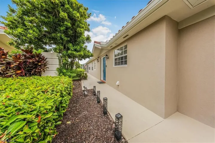 Stucco exterior with a tile roof, concrete walkway, and established landscaping featuring mature trees and shrubs