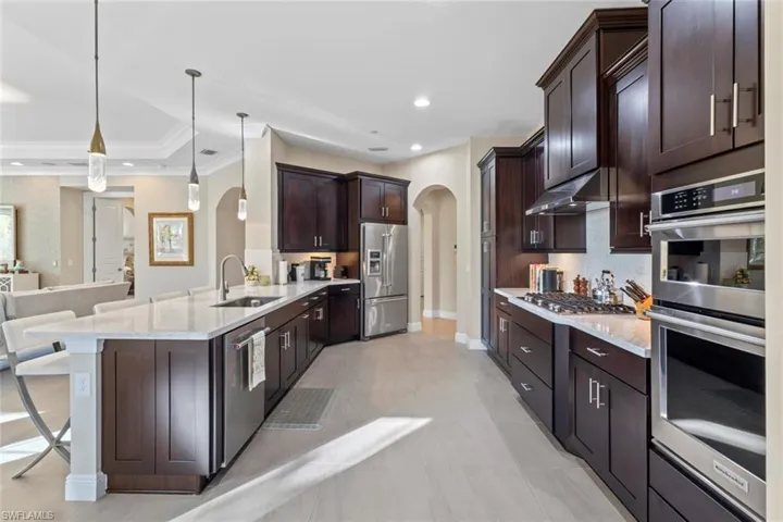 Kitchen with arched walkways, appliances with stainless steel finishes, pendant lighting, light stone countertops, and dark brown cabinetry