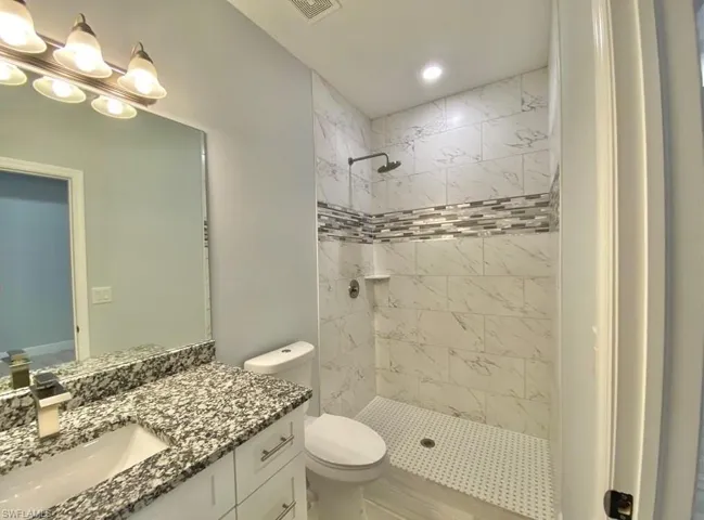 Shower with white subway tile and decorative mosaic band, white vanity with granite countertop and integrated sink, wall-mounted mirror, and multi-bulb vanity light fixture