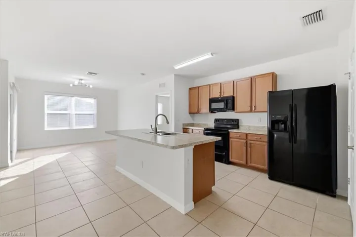 Open-concept living space featuring a kitchen island with a built-in sink, black appliances, wood-finish cabinetry, and tiled flooring