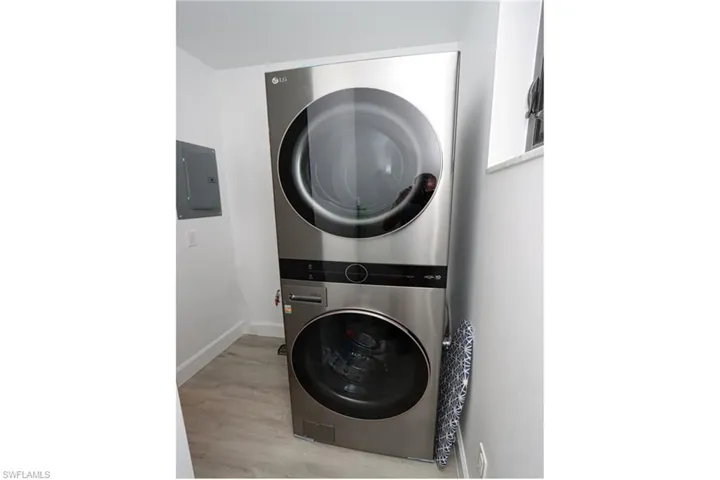 Laundry room with stacked washer and clothes dryer, electric panel, light wood-type flooring, and baseboards
