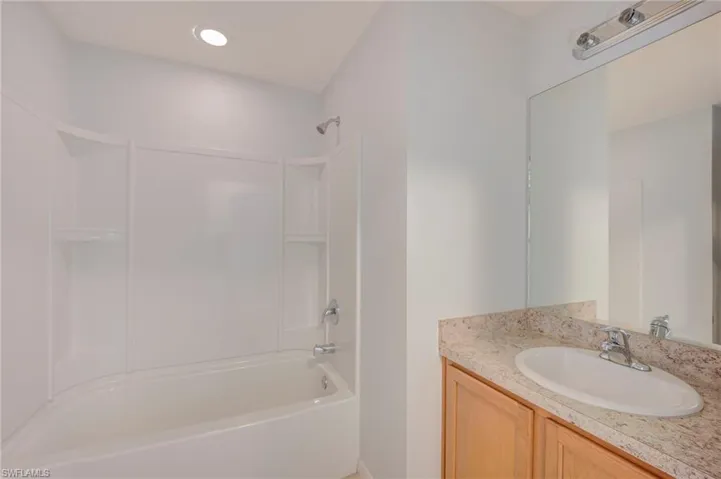 Full bathroom featuring an integrated tub and shower surround, stone-composite vanity countertop with an inset oval sink, wooden cabinetry, and wall-mounted vanity lighting
