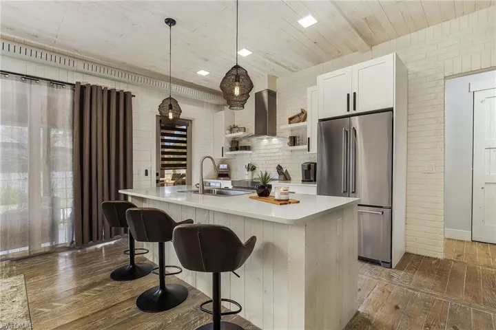 Kitchen with wall chimney range hood, white cabinets, an island with sink, stainless steel fridge, and sink