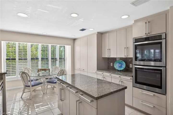 Recessed lighting illuminates a kitchen featuring a large window with white plantation shutters, an expansive central island with a granite countertop, light-toned cabinetry, and stainless steel double wall ovens