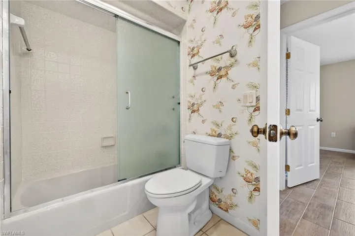 Full bathroom featuring a white bathtub with a sliding frosted glass enclosure, tiled shower surround, a white toilet, and decorative wallpaper