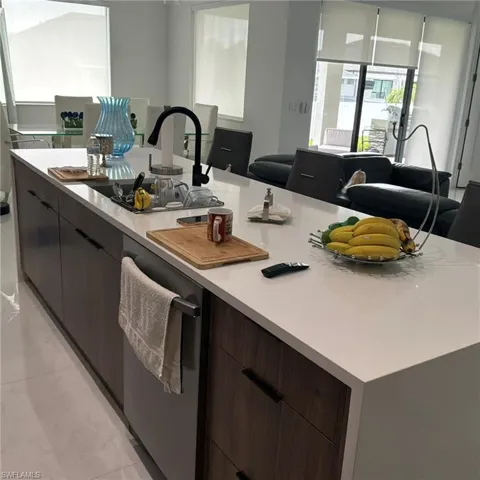 Expansive kitchen island featuring a white countertop, integrated sink with a matte black faucet, wood-finish cabinetry, and a built-in stainless steel dishwasher
