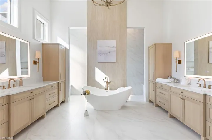Full bath with two vanities, a high ceiling, a freestanding bath, light marble finish floors, and suspended lighting