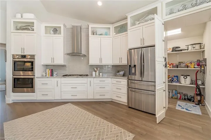 Kitchen with large hidden pantry, wall chimney range hood, light stone countertops, stainless steel appliances, and wood-type flooring