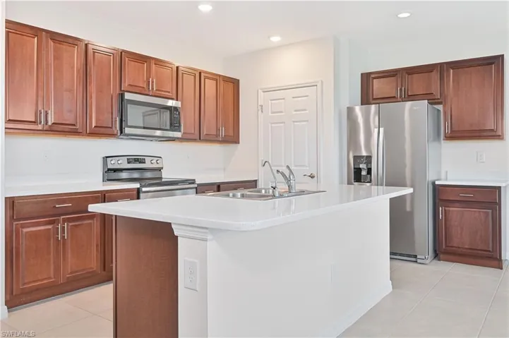 Kitchen with light tile patterned floors, stainless steel appliances, recessed lighting, a center island with sink, and brown cabinetry