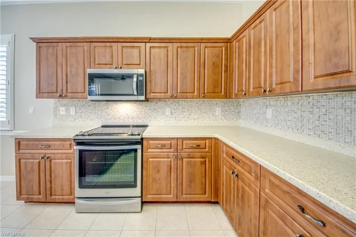 Kitchen with stainless steel appliances, wood finish cabinets, backsplash, light tile patterned floors, and light stone counters