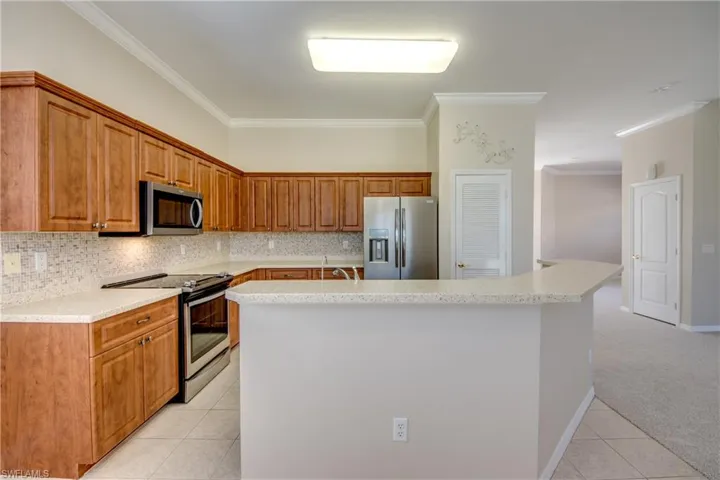 Kitchen featuring stainless steel appliances, light colored carpet, tasteful backsplash, a center island with sink, and crown molding