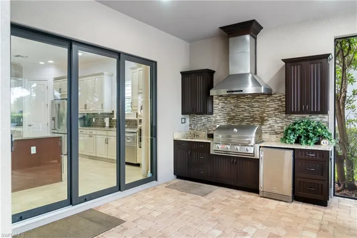 Outdoor kitchen featuring built-in grill, stainless steel range hood, stone tile backsplash, wood-finish cabinetry, and light-toned countertop