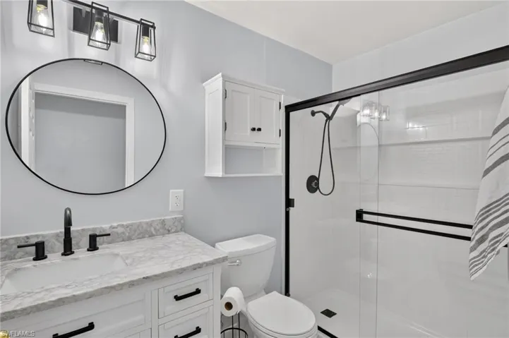 White vanity with a stone countertop, circular mirror, and black-framed shower enclosure