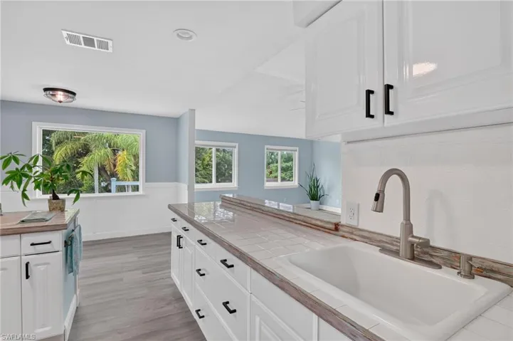 White cabinetry with black hardware, tile countertops, undermount sink with a gooseneck faucet, light wood-finish flooring, and multiple windows