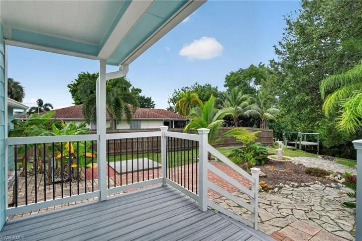 Covered porch with composite decking and white railing, leading to a landscaped yard with stone and brick pathways and mature tropical plantings