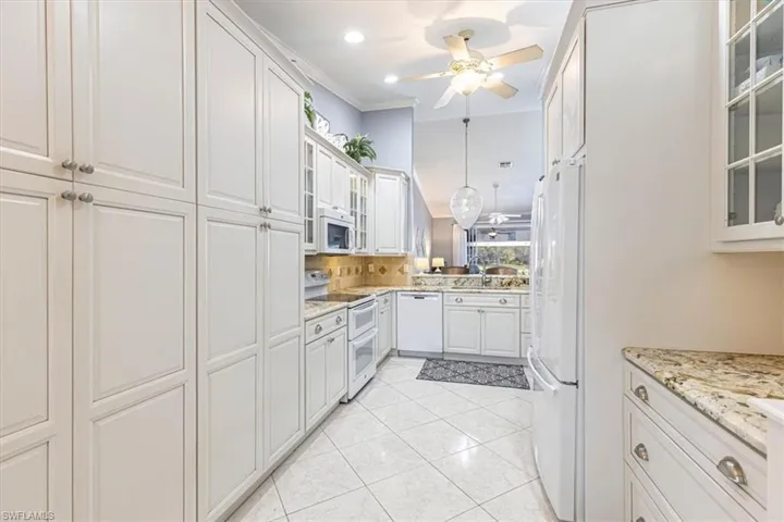 Kitchen w/ glass insert cabinets, white appliances, white cabinetry, light stone countertops & ornamental molding.