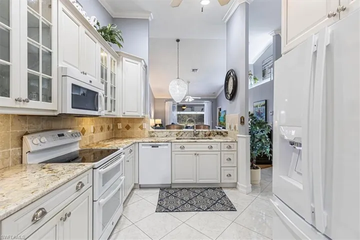 Kitchen featuring white appliances, light stone countertops, decorative backsplash, glass insert cabinets & decorative light fixtures.