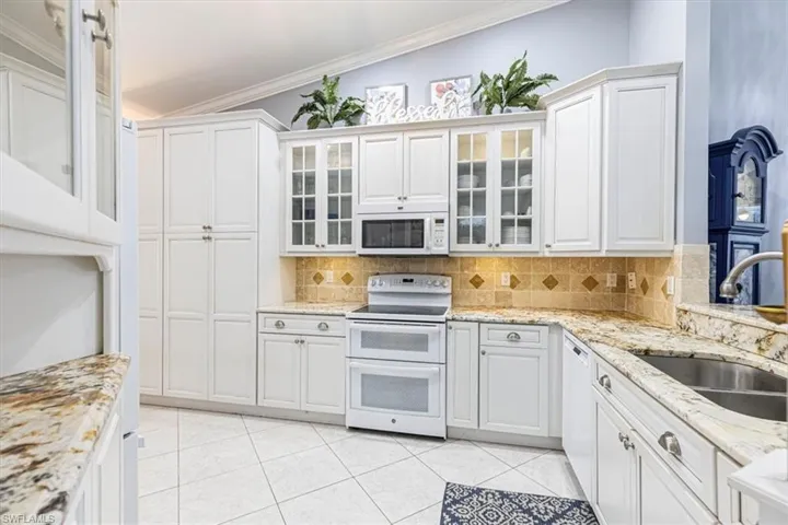 Kitchen with white appliances, light stone counters, white cabinetry, ornamental molding & glass insert cabinets.