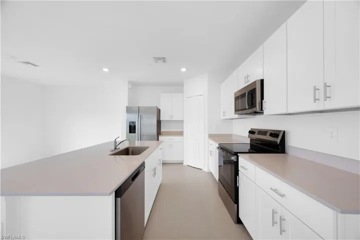 Modern kitchen featuring an expansive center island with an integrated sink and dishwasher, white cabinetry with silver hardware, stainless steel appliances including a refrigerator, microwave, and range, and light-toned countertops