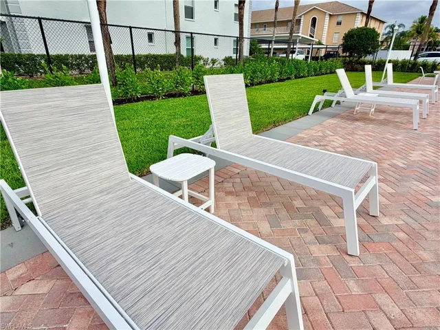 Outdoor lounge area featuring brick paver flooring, multiple lounge chairs with white frames, a white side table, manicured green lawn, and shrubbery