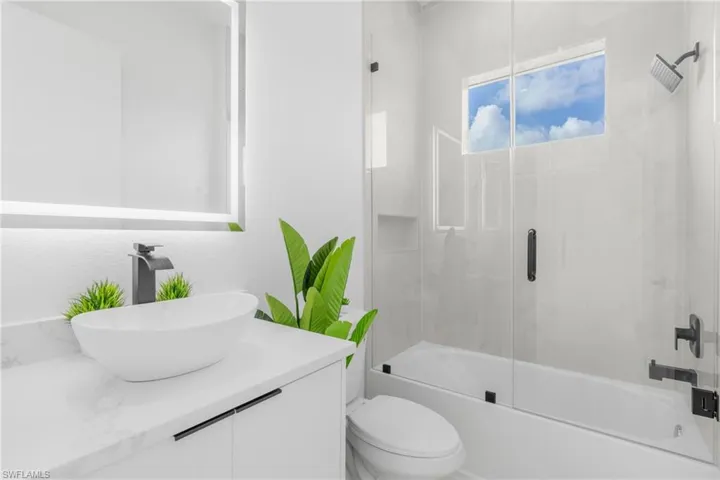 Contemporary bathroom featuring an illuminated vanity mirror, a vessel sink with a matte black faucet, a white countertop, and a glass-enclosed shower over a bathtub with matte black fixtures