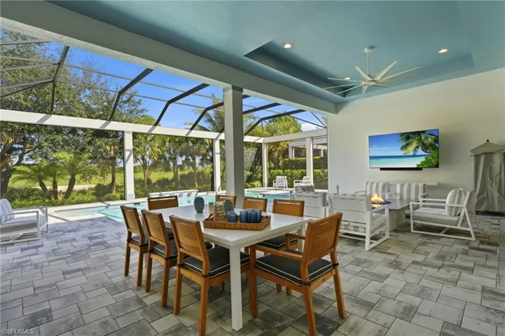 Screened-in outdoor living area featuring a paved patio, recessed tray ceiling with a ceiling fan, and built-in wall-mounted television, overlooking a swimming pool and landscape
