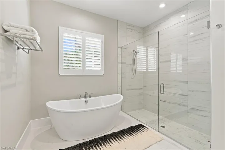 Freestanding soaking tub with a chrome faucet, frameless glass-enclosed shower, floor-to-ceiling tiled shower surround, recessed lighting, and plantation shutters