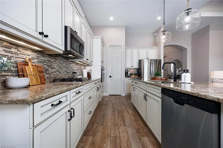 Gourmet kitchen featuring wood-finish flooring, white cabinetry with dark hardware, a mosaic tile backsplash, stainless steel appliances, and a spacious island with a built-in sink and pendant lighting