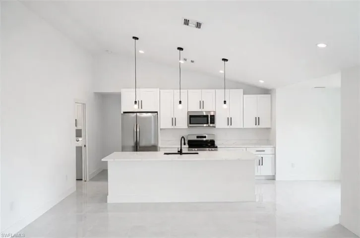 Kitchen featuring stainless steel appliances, white cabinets, a center island with sink, light stone countertops, and vaulted ceiling