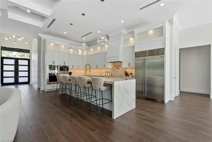 Kitchen with white cabinetry, custom exhaust hood, light stone counters, stainless steel appliances, and a center island with sink