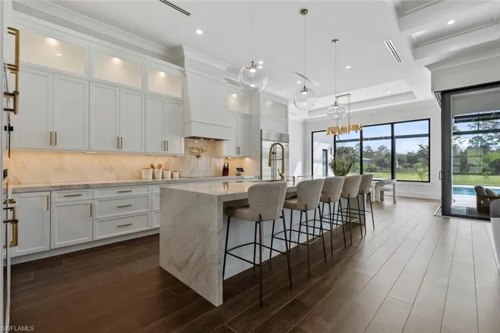Kitchen featuring light stone counters, a center island with sink, white cabinets, pendant lighting, and backsplash