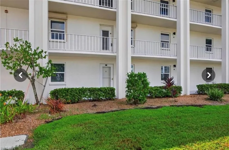 Three-story building exterior featuring white stucco facade, stacked balconies with white railings, and manicured landscaping