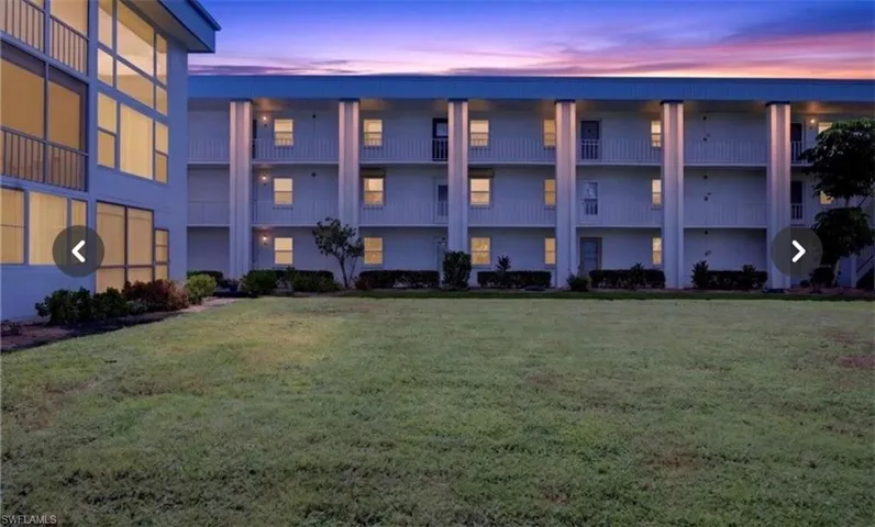 Three-story building exterior featuring vertical columns, stacked balconies, and expansive lawn area