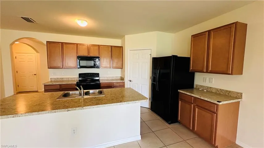 Kitchen with arched walkways, black appliances, brown cabinets, light tile patterned flooring, and light countertops