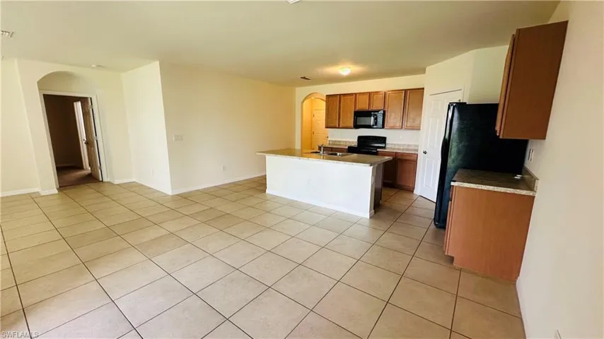 Kitchen featuring arched walkways, black appliances, light tile patterned floors, an island with sink, and brown cabinets