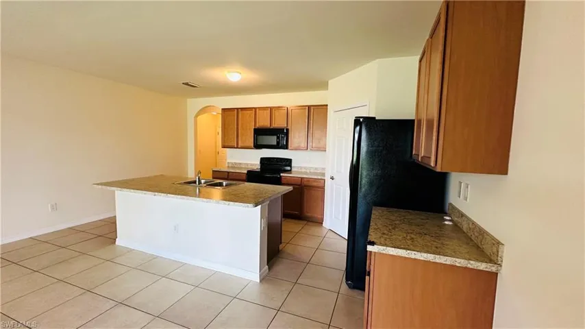Kitchen featuring arched walkways, black appliances, an island with sink, light tile patterned floors, and brown cabinets