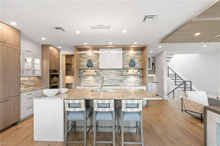 Kitchen featuring light stone counters, pendant lighting, open shelves, a center island with sink, and a breakfast bar area