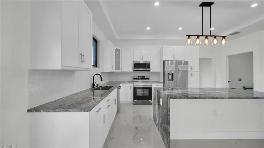 Kitchen featuring dark stone counters, appliances with stainless steel finishes, light marble finish floors, pendant lighting, and white cabinetry